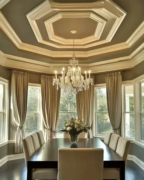 Elegant dining room showcasing a multi-layered tray ceiling with intricate molding, enhanced by a stunning crystal chandelier, neutral decor, and large windows for natural light.