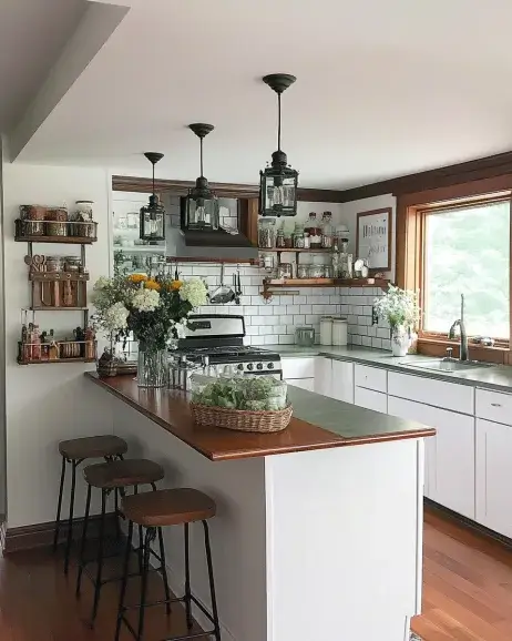 A cozy kitchen decor design featuring white cabinets, a wooden countertop, bar stools, subway tile backsplash, open shelving, and lantern-style pendant lights. The space is accented with fresh flowers and rustic decor elements.